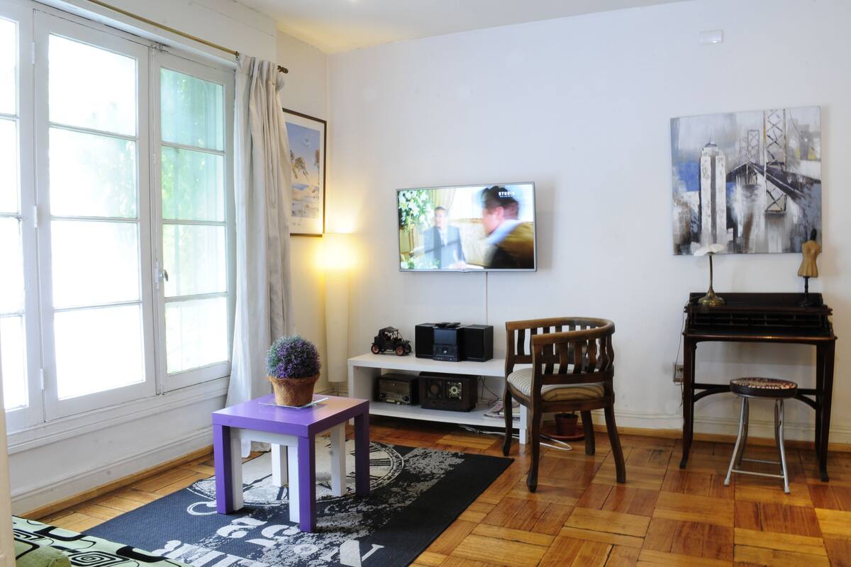 Een woonkamer met een televisie, een piano, een houten stoel, een kleine tafel met een plant en een raam met gordijnen.