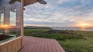 Eine Holzterrasse mit Glaswand, die auf eine Wiese und den Ozean im Sonnenuntergang blickt.