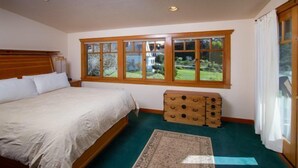 A bedroom with a large bed, a wooden chest of drawers, a window with a view of greenery, and a patterned rug. - Mendocino View East (Mendocino)