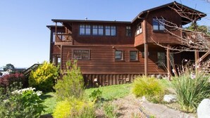 A two-story wooden house with a balcony on the upper floor, surrounded by a landscaped garden with various plants and a pathway. - Mendocino View West (Mendocino)