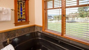 A bathroom with a black bathtub, a stained glass window, and a view of a garden through a window with blinds. - Mendocino View West (Mendocino)