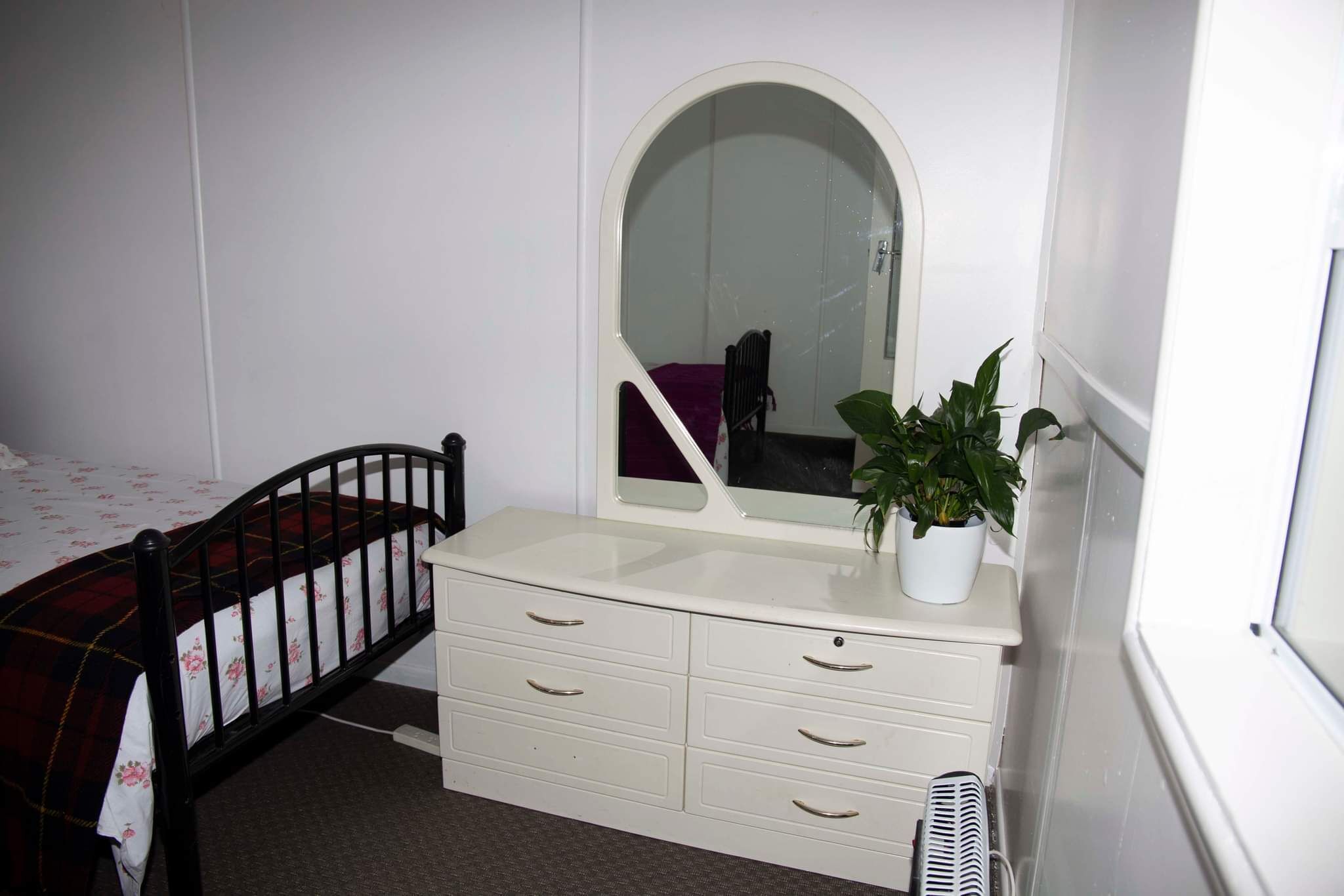 A bedroom with a bed, a dresser with drawers, a mirror, and a potted plant.