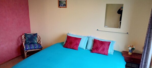 A room with a bed, a chair, a small table with a vase, and a mirror on the wall.