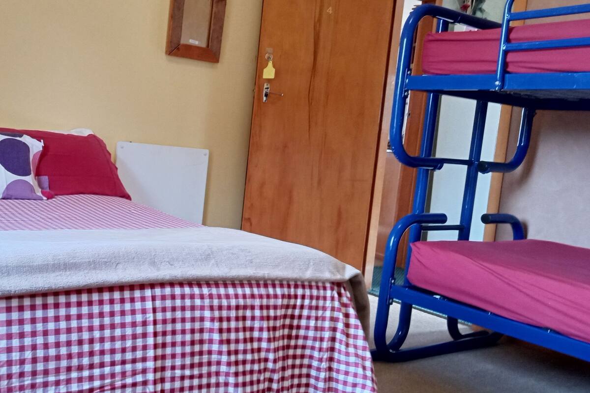 Un lit avec une couvre-lit à carreaux et des oreillers, un lit superposé sur la droite et une porte au fond.