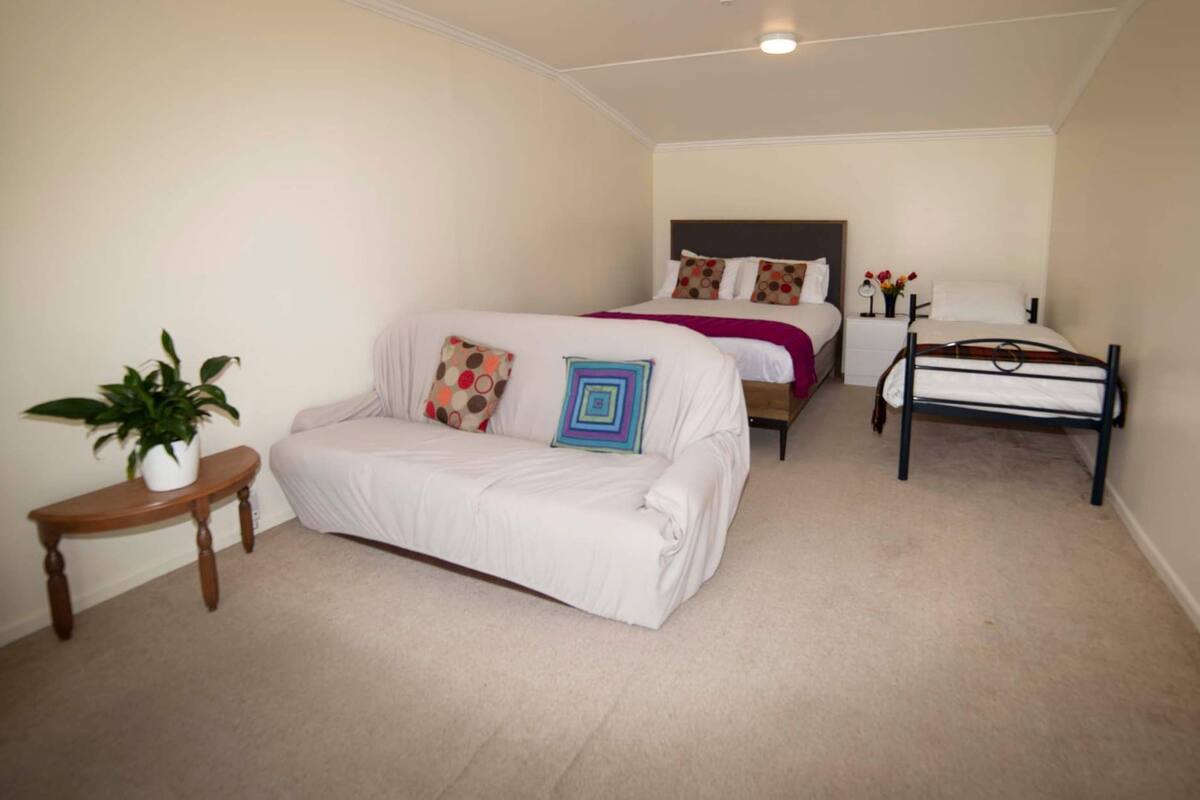 Une chambre avec un canapé blanc, une petite table d’appoint en bois sur laquelle est posée une plante en pot, un lit avec une tête de lit et une table de chevet avec une lampe et des fleurs.