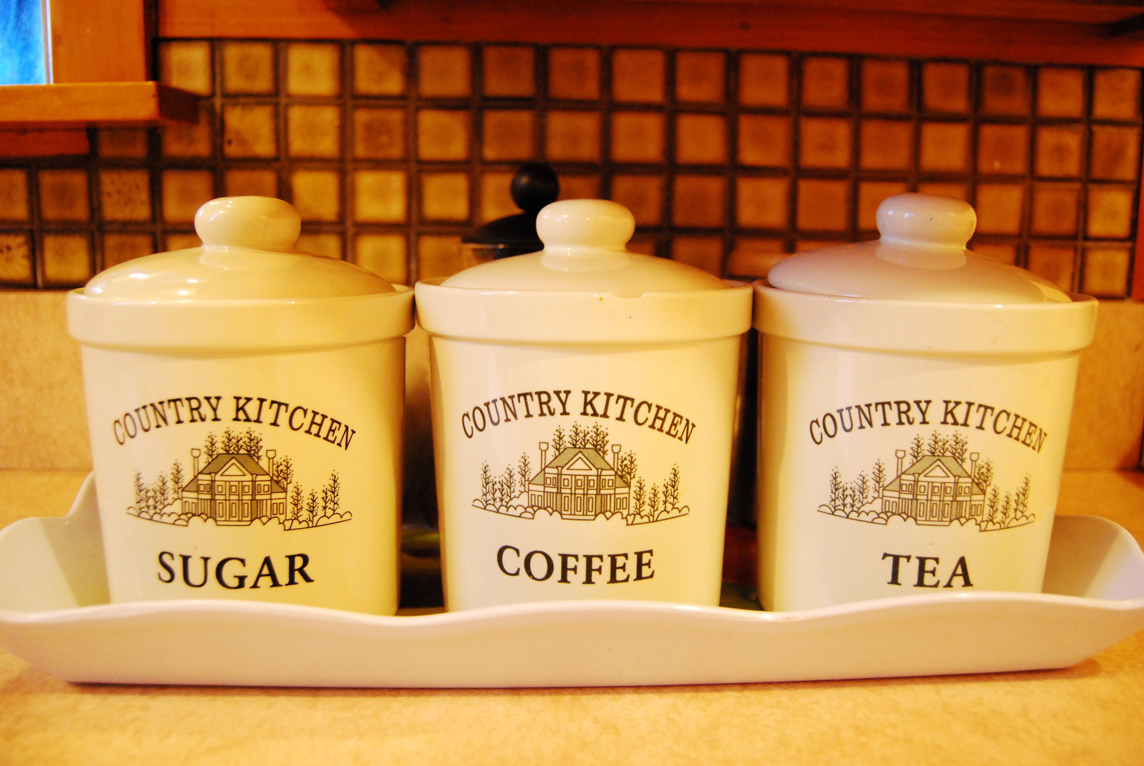Tres recipientes blancos con tapas, etiquetados como 'Country Kitchen Sugar', 'Café' y 'Té', sobre una bandeja.