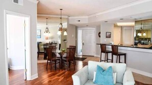 A spacious living area with a dining table and chairs, a sofa with a blue pillow, and a kitchen area with a counter and hanging lights.