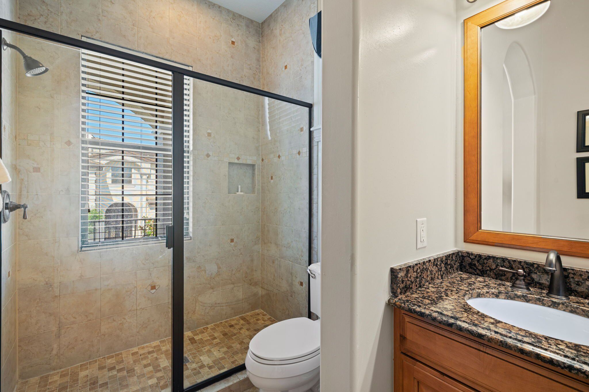 A bathroom with a glass-enclosed shower, a toilet, and a granite countertop sink with a mirror.