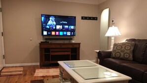 A living room with a flat-screen TV displaying a streaming service interface, a glass-top coffee table, a brown leather sofa, and a lamp. - Awesome remodeled house! (Mentor)
