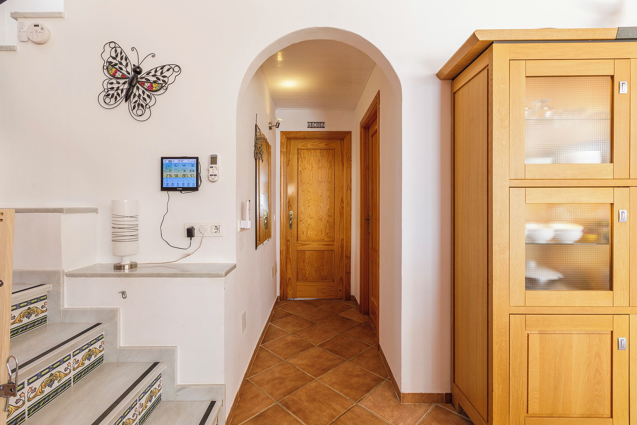 A hallway with a wooden door, a butterfly wall decoration, a digital clock, and a cabinet with glass doors.