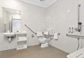 Un baño moderno con lavamanos, inodoro y ducha blancos. Cuenta con barras de apoyo, un perchero y un estante con artículos de aseo.