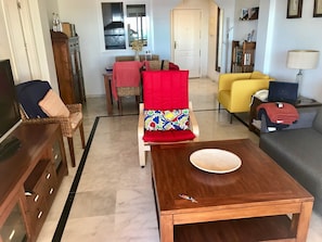 A living room with a wooden coffee table, a sofa, a yellow armchair, and a red chair with a colorful cushion. There is a television, a lamp, and a window with blinds.