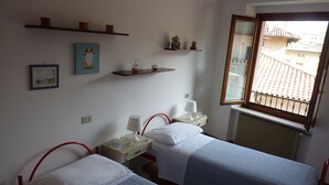 A room with two beds, a window with a view of buildings, and shelves with decorative items.