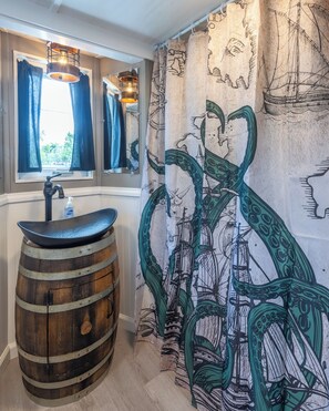 Ein Badezimmer mit einem Waschbecken aus Holzfässern, einem Fenster mit Vorhängen und einer Wand mit einem Seefahrer-Mural.