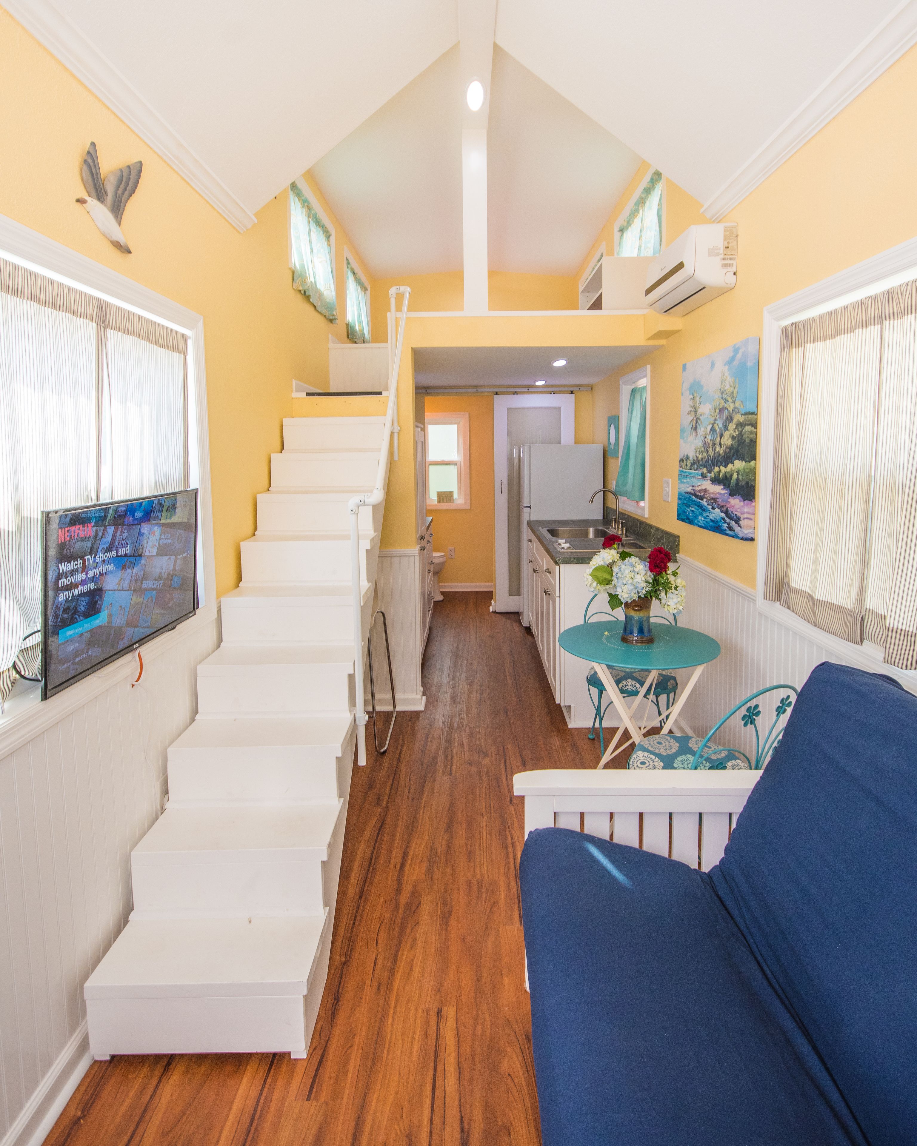 A staircase leading to a kitchen area with a dining table and chairs, a sofa, and a TV mounted on the wall.