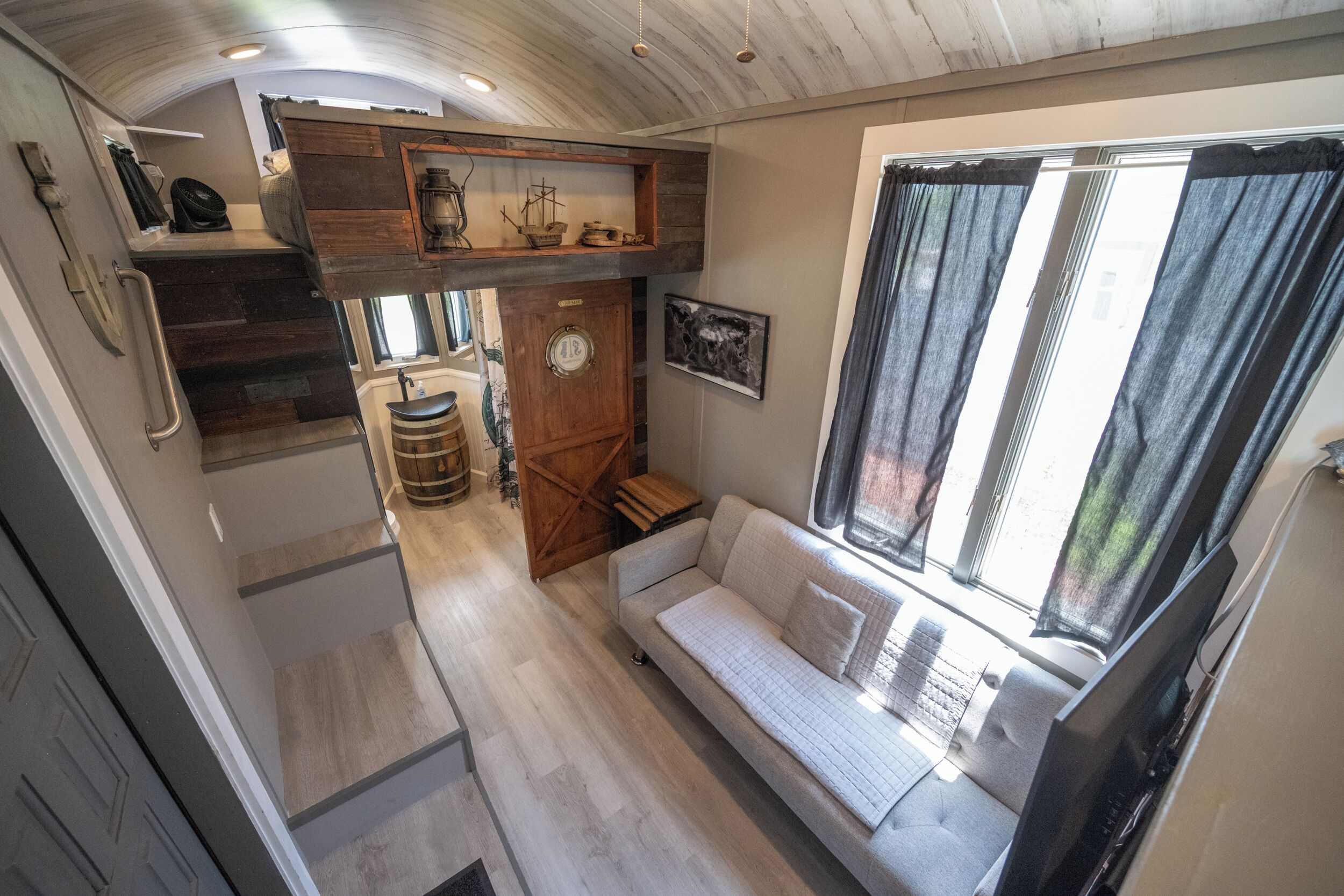 En kompakt opholdsstue med sofa, træhylder, et lille køkken og et vindue med gardiner.