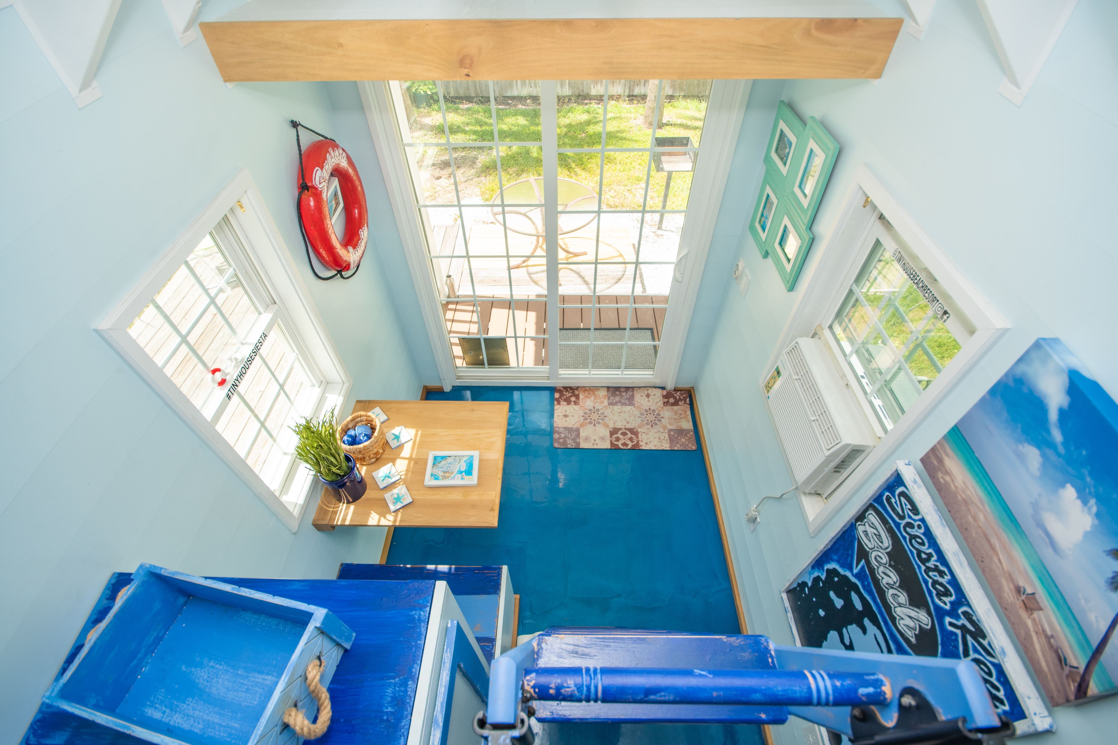 A room with a blue floor, a wooden ceiling, a window with a view of a garden, a red life preserver hanging on the wall, and a sign with a beach scene.