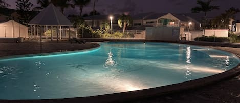 A swimming pool at night with a gazebo and palm trees in the background.