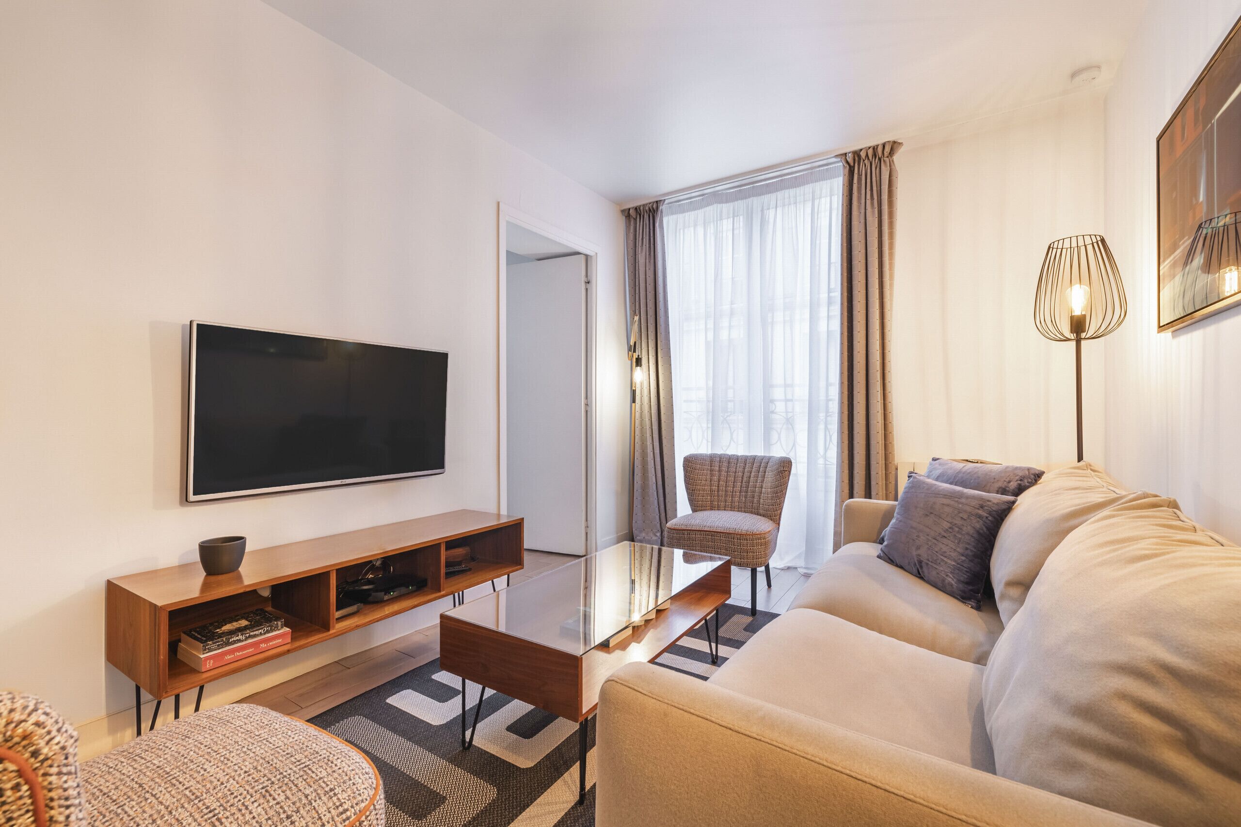 Una sala de estar moderna con una televisión de pantalla plana, una mesa de centro de madera, un sofá color beige, un sillón de mimbre y una lámpara de pie.