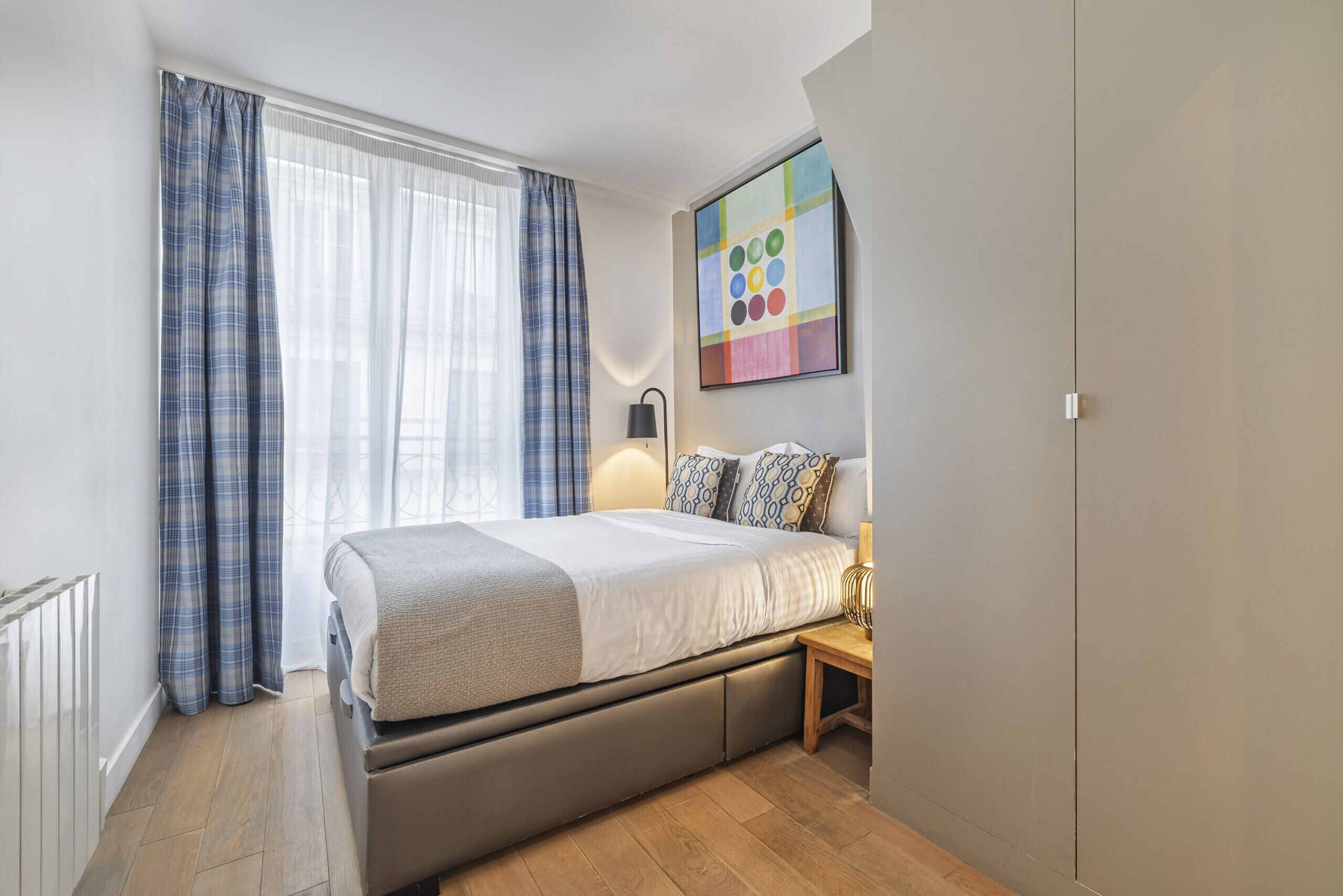 Ein modernes Schlafzimmer mit Bett, Nachttisch, Lampe und einem farbenfrohen, abstrakten Gemälde an der Wand.