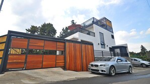 A modern two-story house with a large wooden garage door, a balcony on the upper floor, and two cars parked in front. - NonilSO Private Pension (Seogwipo)