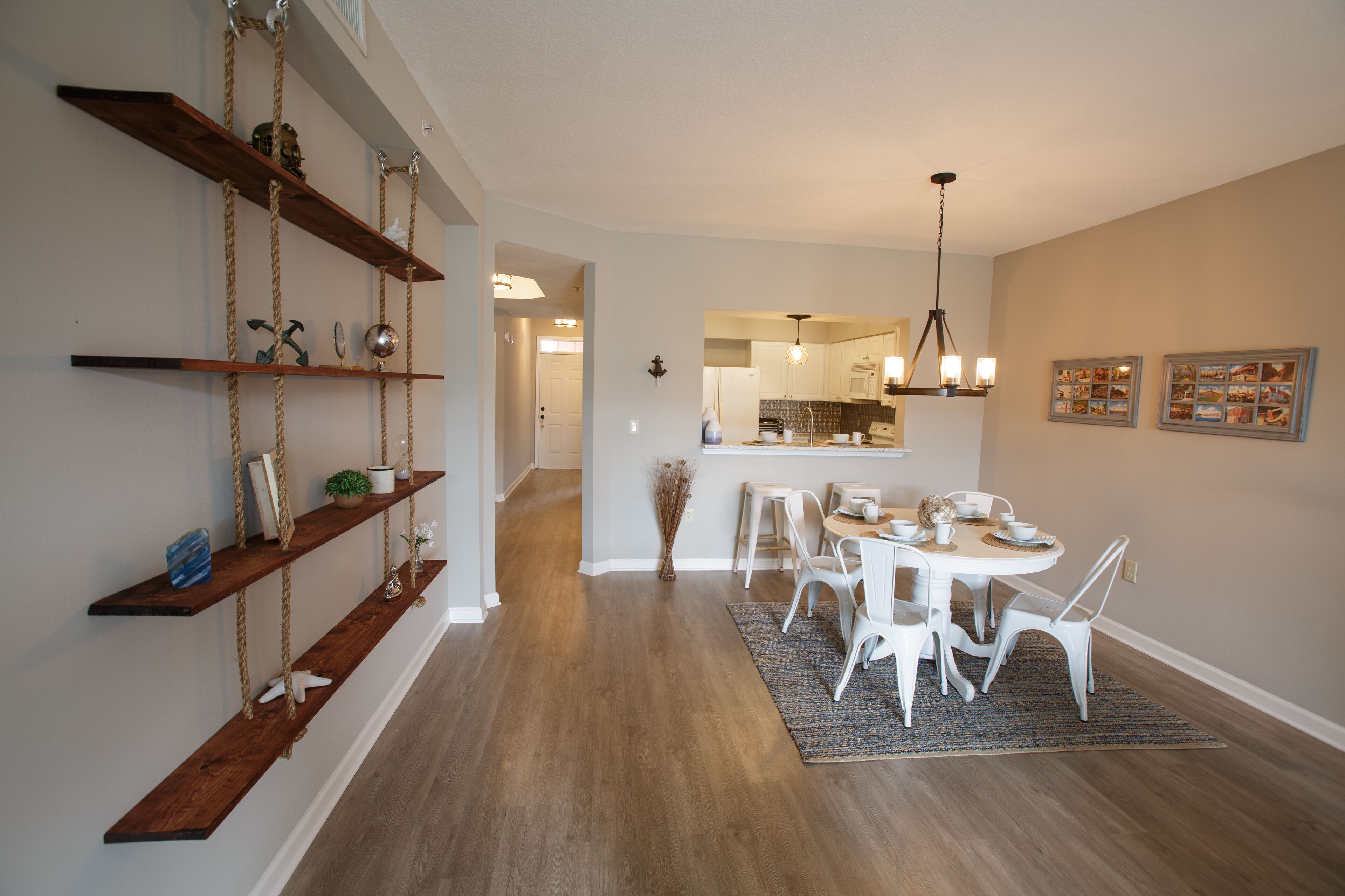 Una cocina moderna con armarios blancos, una zona de comedor con sillas blancas y estantes de madera con objetos decorativos.