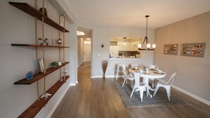 Une cuisine moderne avec des meubles blancs, un coin repas avec des chaises blanches et des étagères en bois ornées d’objets décoratifs.