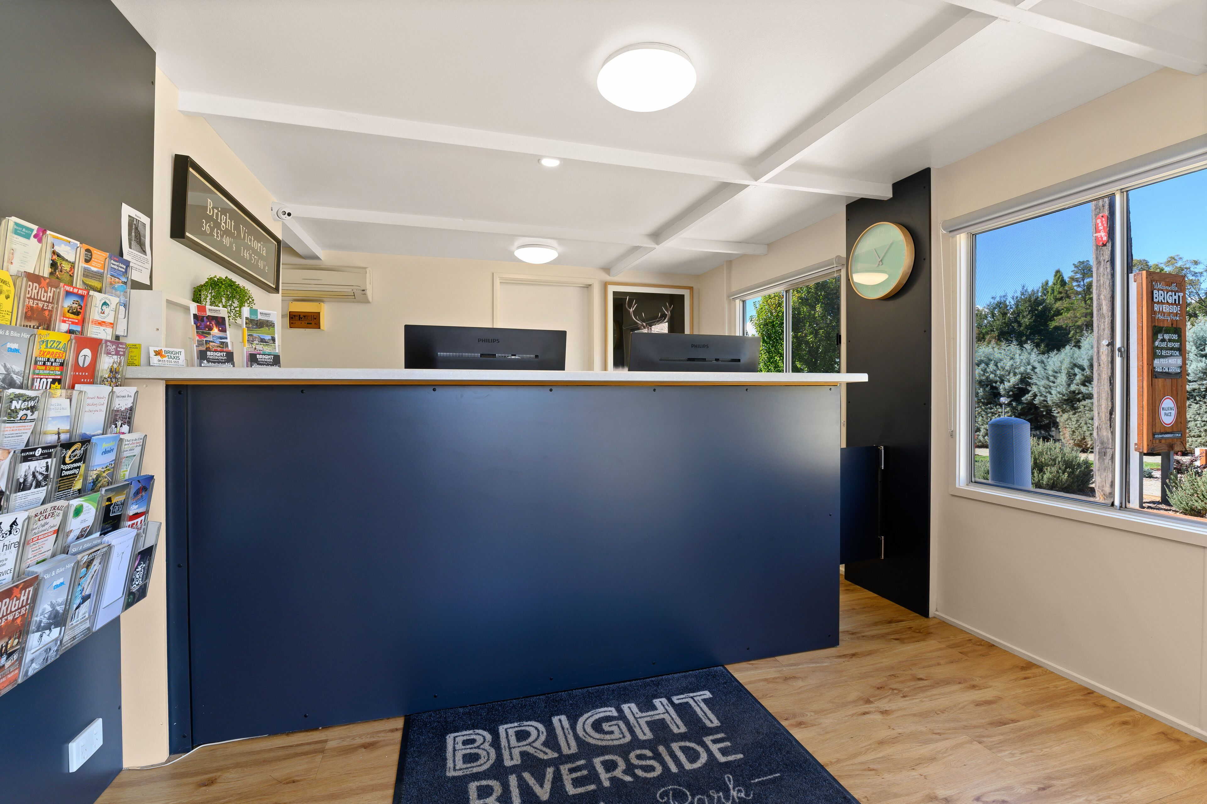 A reception desk with a dark blue counter, a wall-mounted clock, and a window with a view of trees.