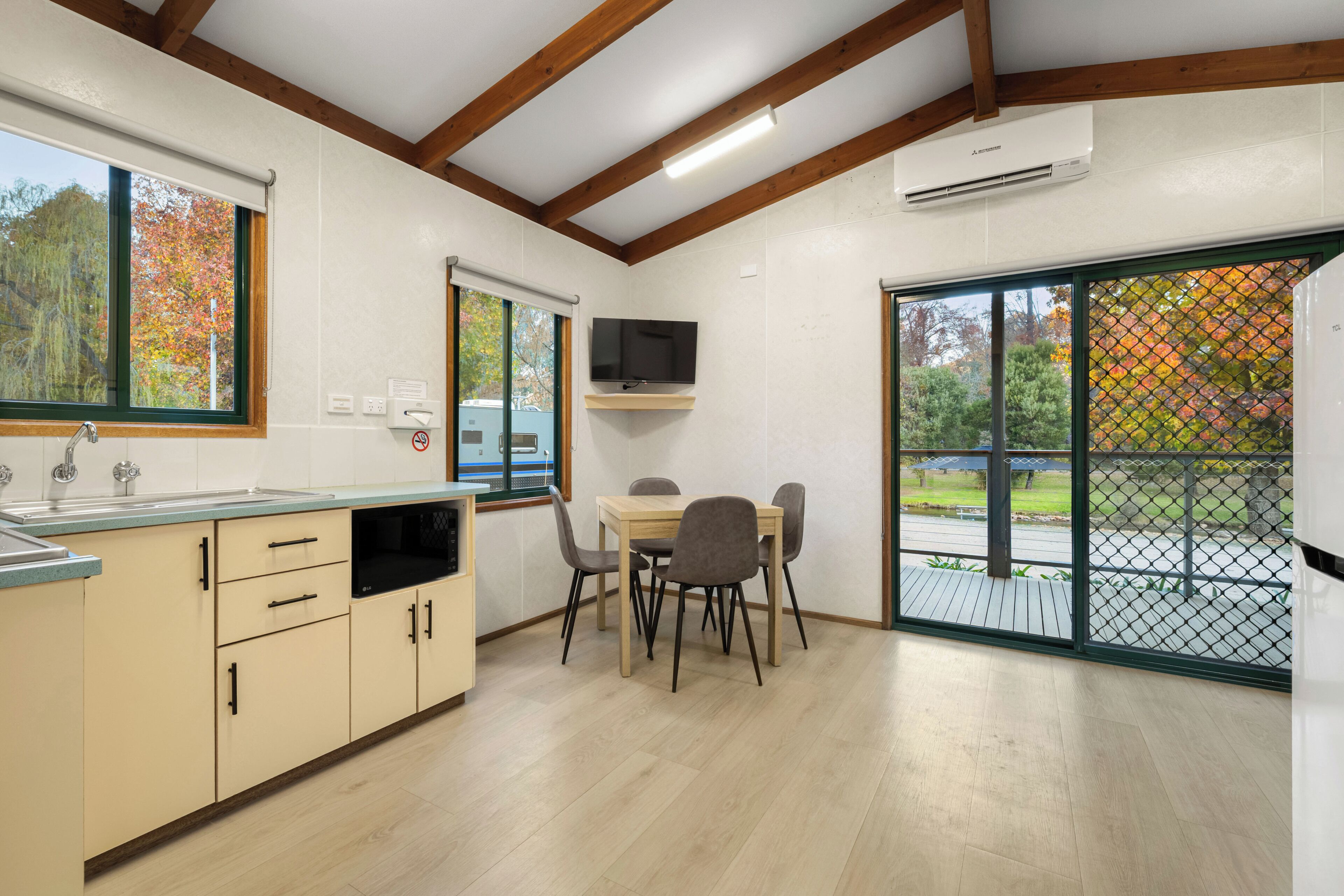 Una cocina moderna con comedor, vigas de madera, televisor y vista a árboles otoñales.