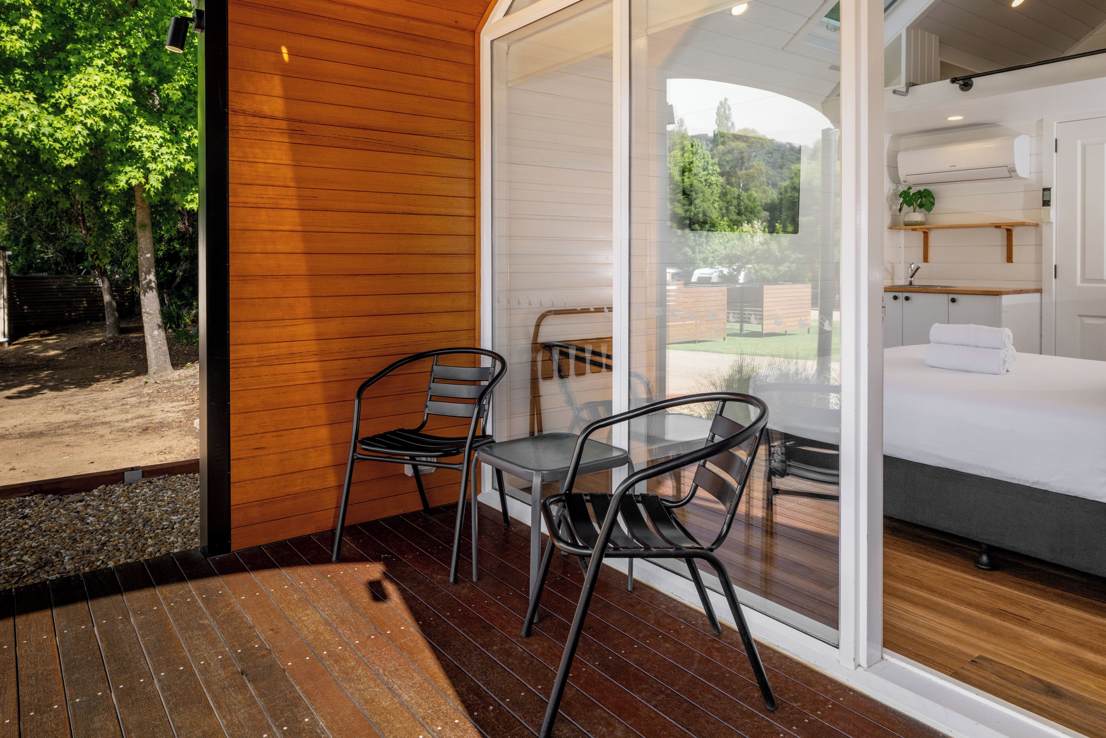 A modern room with a wooden floor, a bed with white linens, a small table with chairs, and a glass door leading to a garden.
