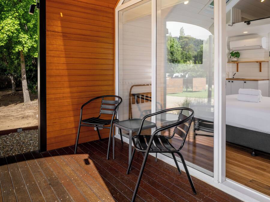 A modern room with a wooden floor, a bed with white linens, a small table with chairs, and a glass door leading to a garden.