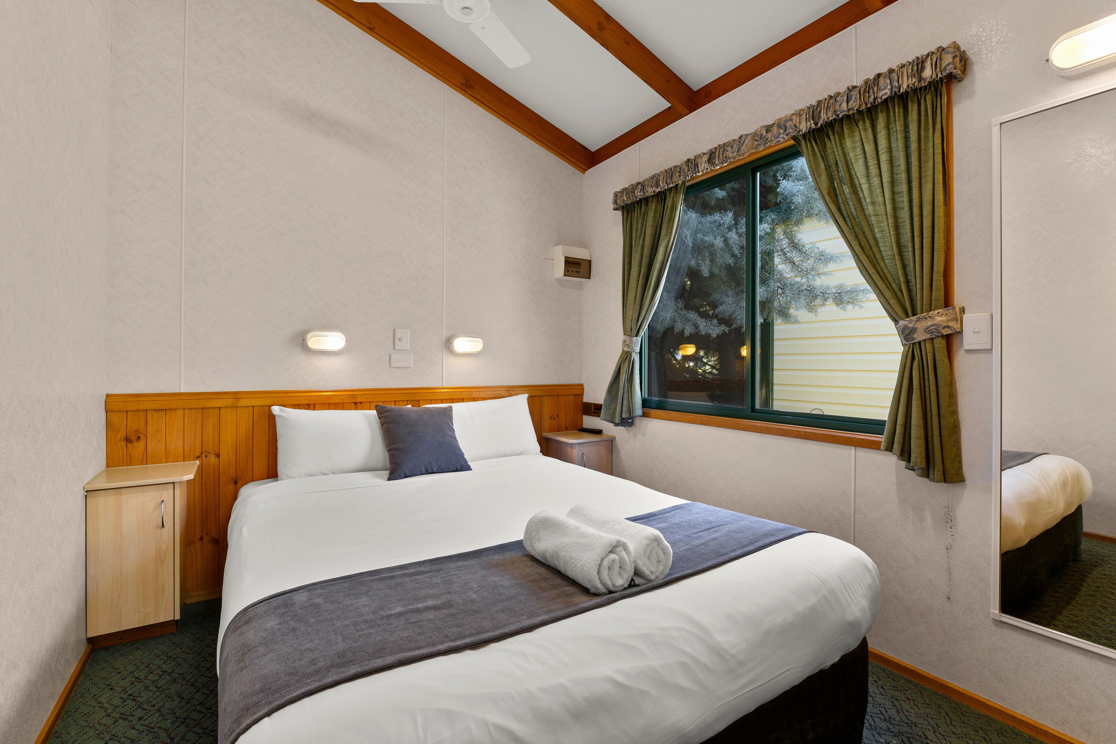 A neatly made bed with a headboard, a bedside table, a nightstand, and a window with curtains.