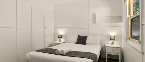A neatly arranged bedroom with a large bed, two bedside tables with lamps, a wardrobe, and a window with a view.