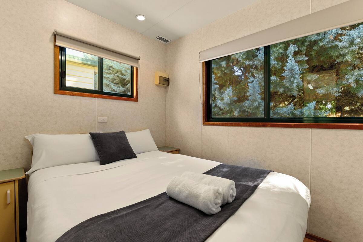 A neatly made bed with white and gray linens, a dark gray throw blanket, and a folded white towel. There is a small bedside table with a lamp and a wall-mounted air conditioning unit. Two windows provide views of trees and a building.