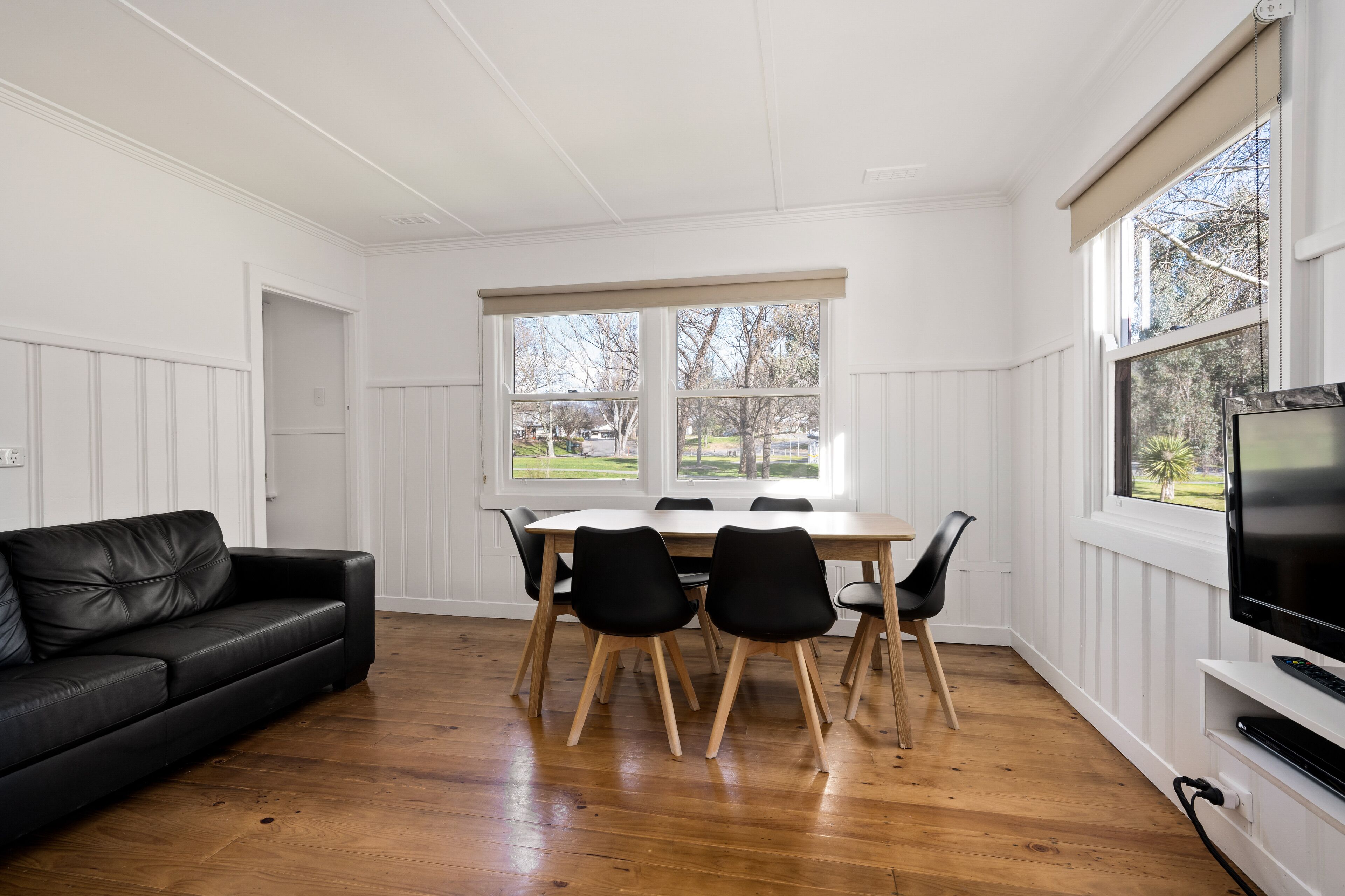 A room with a dining table, chairs, a black leather sofa, a TV, and large windows with a view of the outdoors.