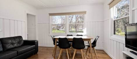 A room with a dining table, chairs, a black leather sofa, a TV, and large windows with a view of the outdoors.