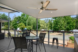 Un espacio al aire libre techado con una mesa, sillas, ventilador de techo y vistas a árboles y vehículos al fondo.