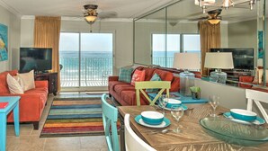 A modern living room with a large TV, a dining area with a table set for two, and a balcony with a view of the ocean. - Summerlin-Beachfront for Late Summer!  5 Star! (Fort Walton Beach)