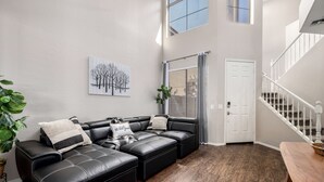 Ein modernes Wohnzimmer mit Ecksofa, großem Fenster mit Jalousien, einer Treppe und einer Tür.