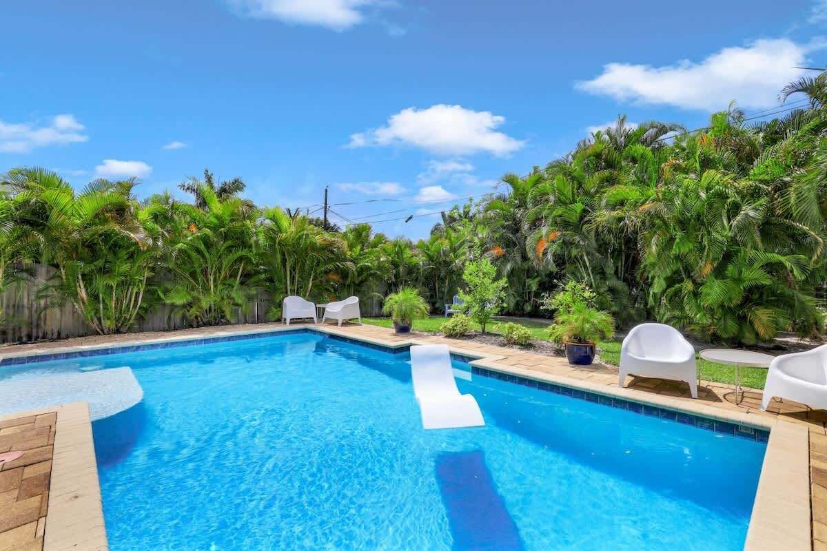 Una piscina con tobogán, rodeada de exuberante follaje y sillas de descanso blancas.