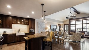 A spacious kitchen with dark wood cabinets, a granite countertop, and a large island. There is a dining area with a table and chairs, and a large window providing a view of the outdoors. - Luxury Ski-In, Ski-Out Arrowleaf Penthouse  With Panoramic Views (Park City)