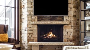 A stone fireplace with a TV mounted above it, a comfortable seating area with a sofa and armchair, and a bookshelf to the right. - Luxury Ski-In, Ski-Out Arrowleaf Penthouse  With Panoramic Views (Park City)