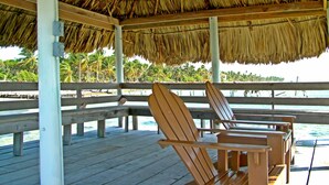 A wooden deck with thatched roof, wooden chairs, and a view of the ocean and palm trees.
