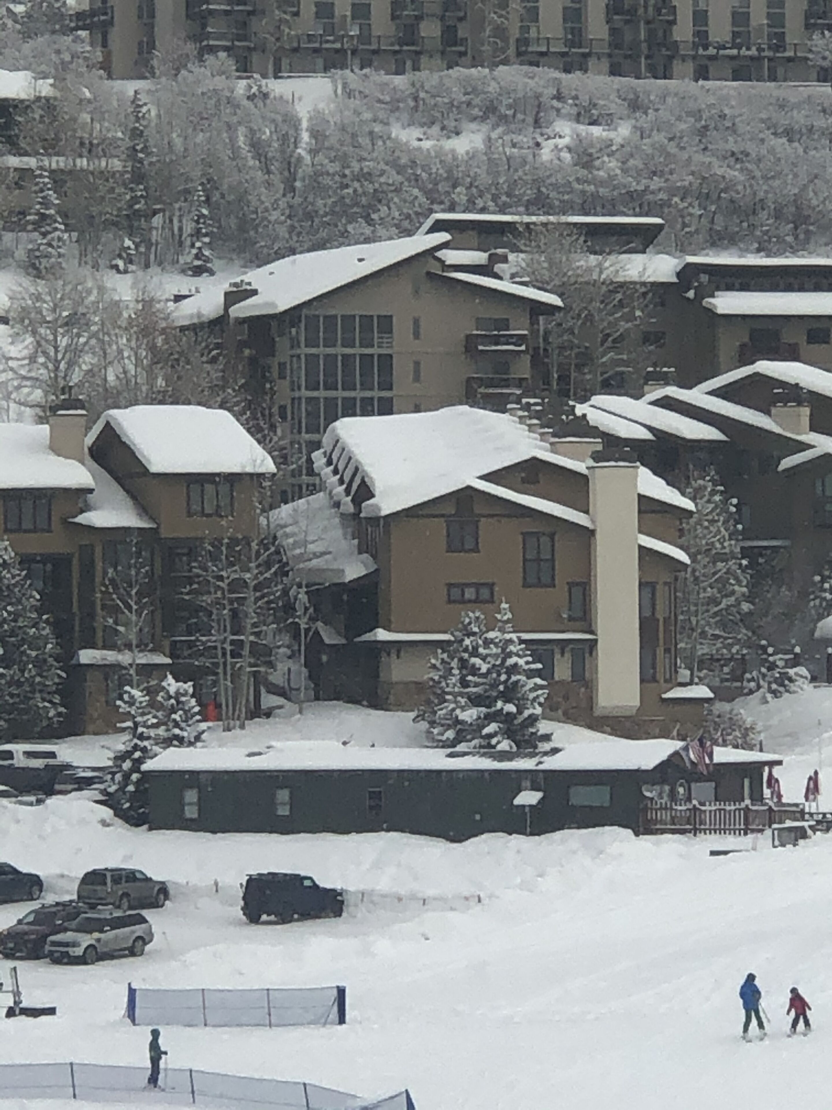 Zona residencial cubierta de nieve con edificios de varios pisos, un estacionamiento con varios autos y algunas personas esquiando.