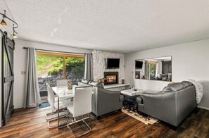 A modern living room with a fireplace, grey sofas, a dining table with chairs, and a large window offering a view of greenery.