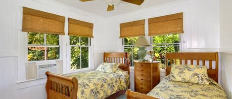 A bedroom with two wooden beds, a ceiling fan, and large windows with bamboo blinds.