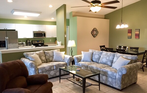 A modern kitchen with stainless steel appliances, a dining area with a glass table, and two patterned sofas.