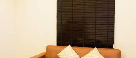A room with a brown sectional sofa, two pillows, a window with blinds, and an air conditioning unit.