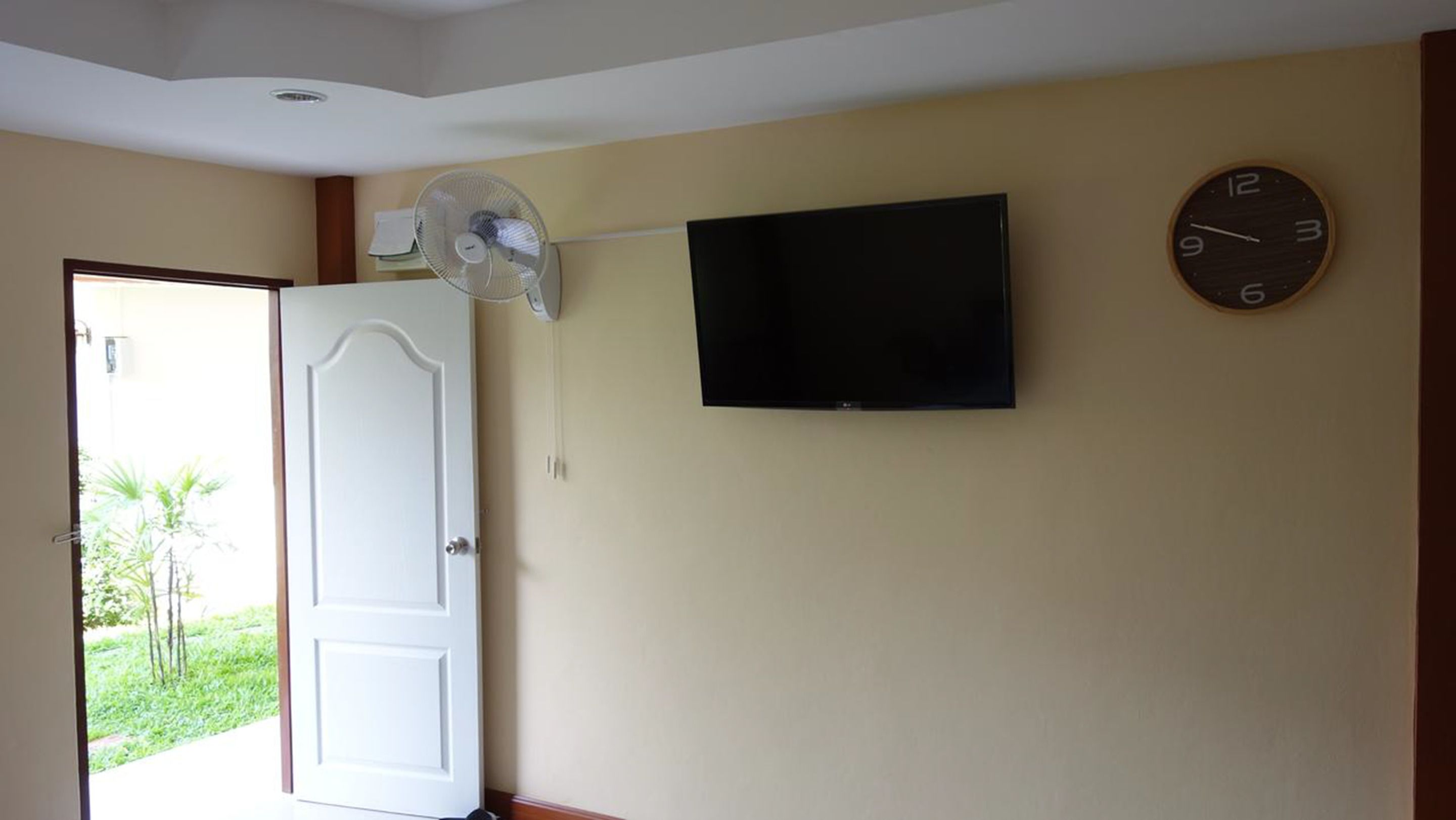 A room with a closed door, a wall-mounted TV, a round clock, and a standing fan.
