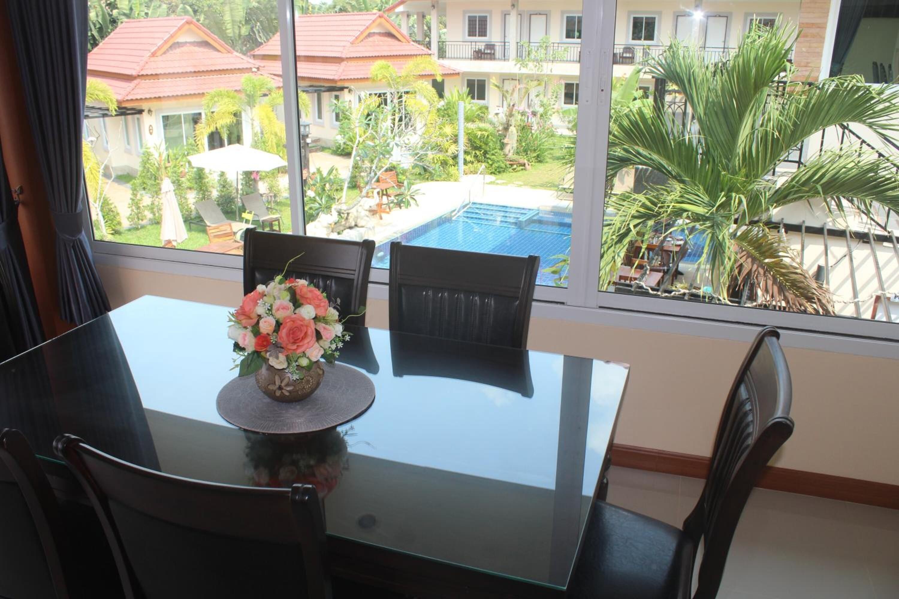 A dining area with a glass table and chairs, a vase of flowers, and a view of a swimming pool and buildings.